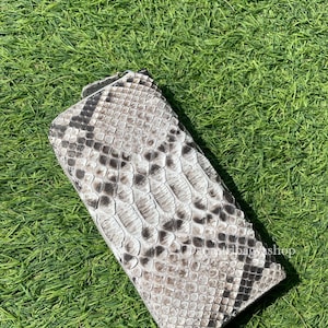 Könnte beinhalten: Eine rechteckige Geldbörse mit Schlangenmuster in Grau- und Schwarztönen. Die Geldbörse liegt auf einem Bett aus grünem Gras, wobei die Textur der Schlangenhaut deutlich sichtbar ist.