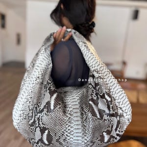 Könnte beinhalten: Eine große Handtasche mit weißem, grauem und braunem Schlangenmuster. Die Tasche hat einen weichen, lässigen Stil mit einer runden Form. Der Text "Danastribagshop" ist auf der Tasche zu sehen.