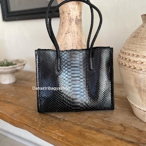 May include: Black rectangular handbag with a snakeskin pattern. The bag has two braided handles and the text "Dahastribagyashop" is visible. The bag is sitting on a wooden surface.