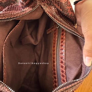 Peut inclure: Vue ouverte d'un sac &agrave; main marron et rouge &agrave; motif peau de serpent. L'int&eacute;rieur est de couleur marron uni, avec une fermeture &eacute;clair et une petite poche zipp&eacute;e. Le texte "Danastribagyashop" est visible.