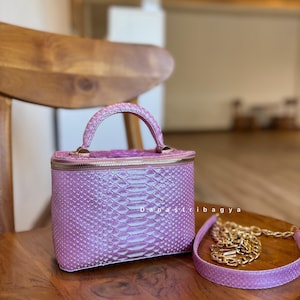 Peut inclure: Un sac à main rectangulaire structuré de couleur lavande avec un motif en peau de serpent texturé, une poignée supérieure et une fermeture éclair dorée. Le sac comprend une bandoulière amovible avec une chaîne dorée. Le texte 'D a n a s t r i b a g y a' est visible.