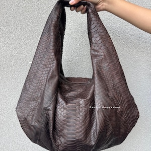 May include: A brown, textured leather shoulder bag with a large, slouchy design. The bag features a wide strap and a snakeskin pattern. The text "Danastribagyashop" is visible on the bag.
