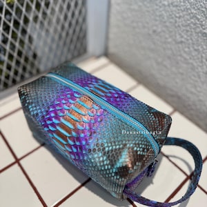 Peut inclure: Une trousse de toilette rectangulaire avec un motif peau de serpent irisé dans des tons bleu métallique, violet et cuivre. Le sac possède une fermeture éclair supérieure avec un liseré bleu clair et une dragonne assortie. Le texte Danastribagya est imprimé sur le côté.