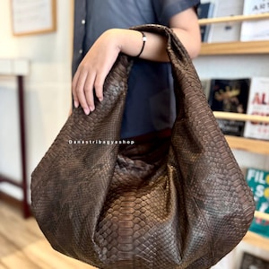 Könnte beinhalten: Eine große, braune, strukturierte Handtasche mit Schlangenmuster. Die Tasche wird von einer Person gehalten. Die Tasche hat eine breite, gebogene Form und einen einzelnen Riemen. Der Text "Danastribagyashop" ist auf der Tasche zu sehen.