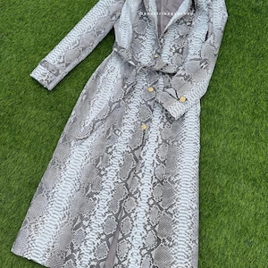 May include: A long, light grey and white snakeskin-patterned coat with gold buttons and a matching belt. The coat is laid out on a green surface. The coat has long sleeves and a collared neckline.