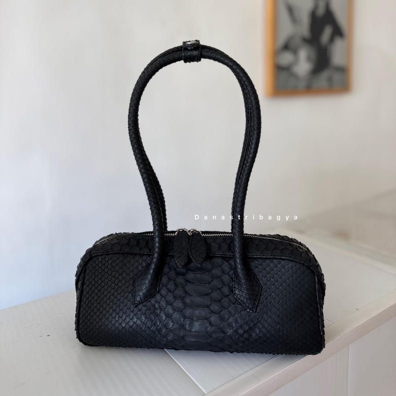 May include: A black handbag with a textured snakeskin pattern, featuring a single arched handle and a top zipper closure with two pulls. The bag has a rectangular, elongated shape and includes the text Danastribagya printed in white on the front.