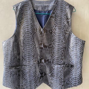 Puede incluir: Un chaleco gris con estampado de piel de serpiente, cuello en V y dos bolsillos delanteros. El chaleco tiene un forro azul oscuro y está colgado de una percha verde. El texto "banastribagyshop" es visible en la parte superior.