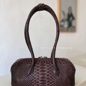 May include: A cylindrical handbag featuring a dark brown snakeskin texture with a repeating scale pattern. The bag has two matching rounded handles and a silver-toned zipper closure along the top. The text Danastribagya is printed in white.
