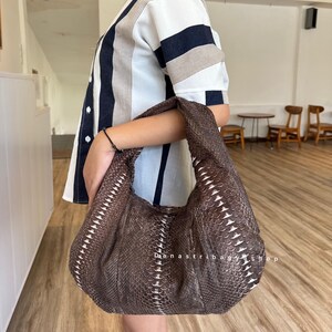Puede incluir: Un bolso marrón texturizado con forma de media luna. El bolso presenta un patrón de escamas con detalles blancos. El texto "Danastribagshop" es visible en el bolso. El bolso lo lleva una persona con una camisa a rayas.