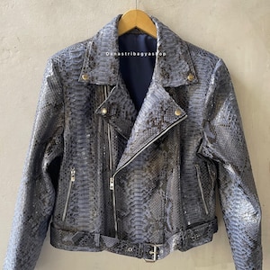 Może przedstawiać: Niebiesko-szara skórzana kurtka ze wzorem skóry węża w klasycznym stylu motocyklowym. Posiada asymetryczny zamek błyskawiczny, klapy z wycięciami i pasek w talii. Kurtka wisi na drewnianym wieszaku.