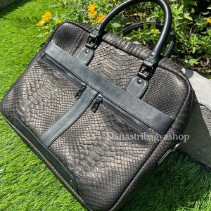 May include: Black leather briefcase with a snakeskin texture, featuring a top handle and a shoulder strap. The bag has two zippered compartments and is set against a backdrop of green grass and yellow flowers. The text "Danastribagyshop" is visible on the bag.