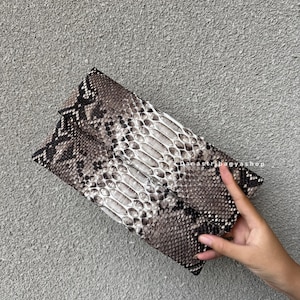 Puede incluir: Un bolso de mano con estampado de piel de serpiente en tonos marrón, gris y blanco. El bolso rectangular tiene una solapa y una superficie texturizada. El bolso se sostiene sobre un fondo gris.