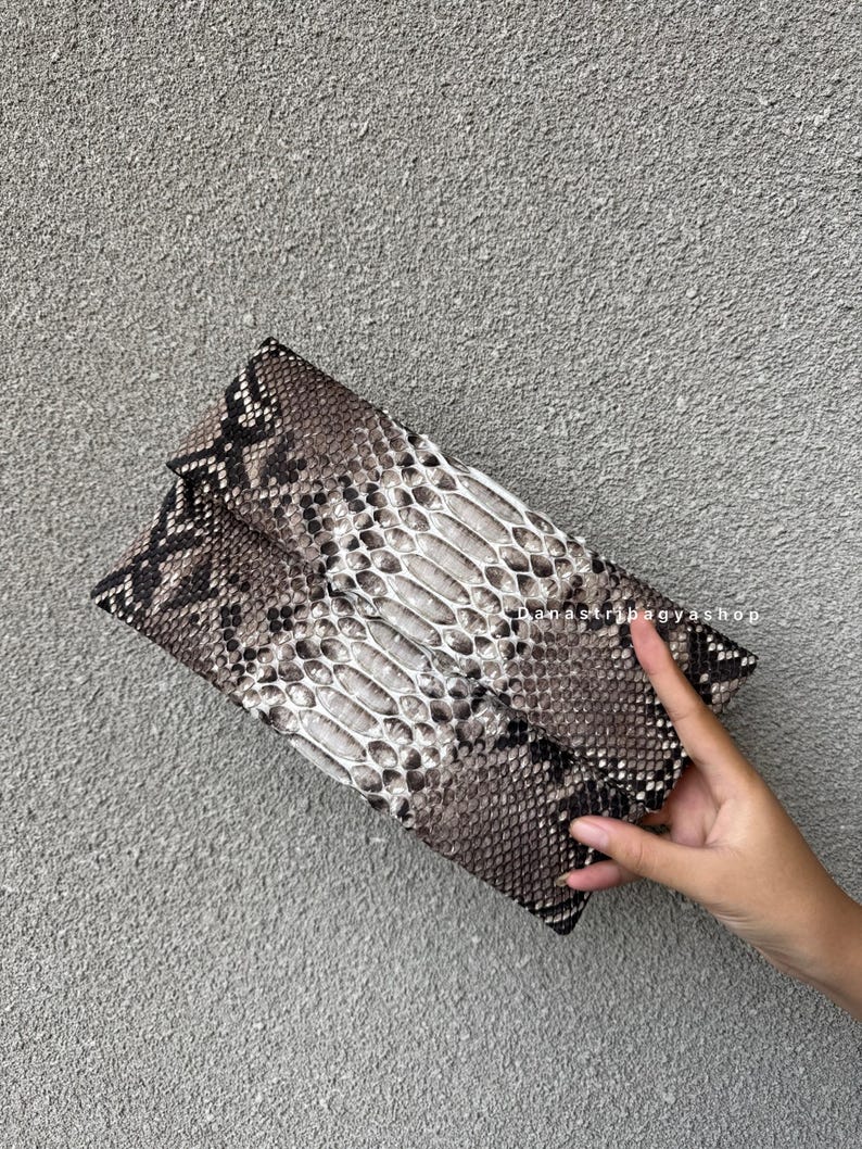 K&ouml;nnte beinhalten: Eine Clutch mit Schlangenmuster in Braun-, Grau- und Wei&szlig;t&ouml;nen. Die rechteckige Tasche hat eine Umschlagklappe und eine strukturierte Oberfl&auml;che. Die Tasche wird vor einem grauen Hintergrund gehalten.