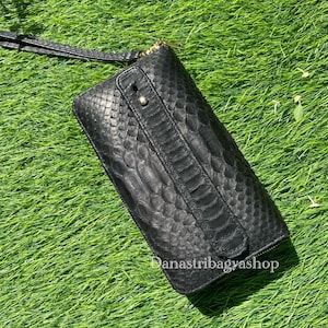 Pode incluir: Bolsa de mão de couro preta com padrão de pele de cobra texturizado, uma alça de pulso e um zíper dourado. A bolsa retangular está sobre a grama verde. O texto "Danastribagyashop" é visível.