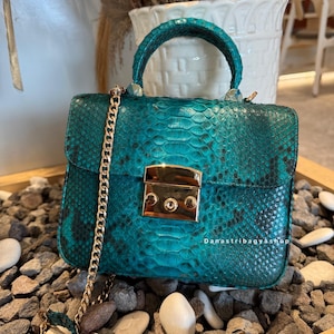 Puede incluir: Un bolso de mano turquesa con estampado de piel de serpiente, con una correa de cadena y un cierre dorados. El bolso tiene un asa superior y forma rectangular. El texto "Damastribagyashop" es visible.