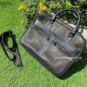 Pode incluir: Pasta preta com textura de pele de cobra, com uma alça superior e uma alça de ombro removível. A bolsa tem um bolso frontal com zíper e formato retangular. A bolsa está sobre a grama verde.