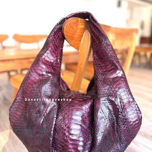May include: A deep purple handbag with a snakeskin pattern. The bag is draped over the back of a wooden chair. The text "Danastri Bagyashop" is visible on the front of the bag.