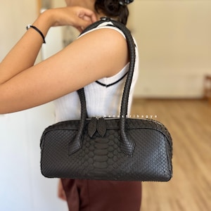 May include: A dark charcoal gray handbag with a textured snakeskin pattern. The bag features a cylindrical shape, two sturdy top handles, and a silver-toned zipper closure with two matching pull tabs. The surface has a distinct scale-like finish.
