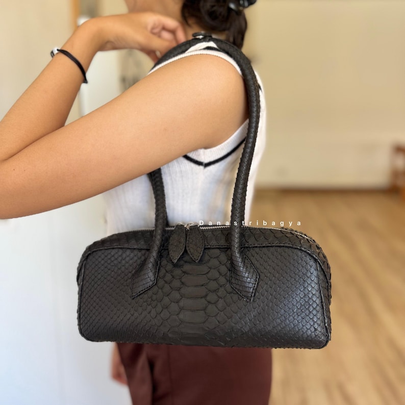 May include: A dark charcoal gray handbag with a textured snakeskin pattern. The bag features a cylindrical shape, two sturdy top handles, and a silver-toned zipper closure with two matching pull tabs. The surface has a distinct scale-like finish.