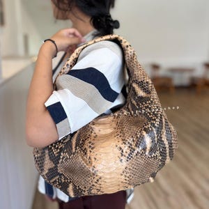 May include: A large, brown and tan snakeskin shoulder bag. The bag has a rounded shape and a single strap. The snakeskin pattern is visible across the entire surface of the bag. The bag is being carried over the shoulder.