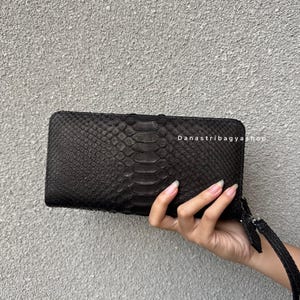May include: A black, rectangular wallet made of textured material, possibly leather or a similar material. The wallet has a zipper closure and a wrist strap. The image includes the text "Danastribagyshop".