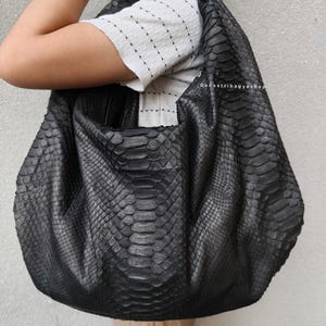 Puede incluir: Un bolso grande de cuero negro con un patrón de piel de serpiente. El bolso es de color oscuro y tiene forma redondeada. El bolso lo sostiene una persona. El texto "Danastribagyashop" es visible en el bolso.