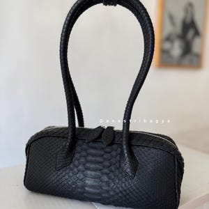 May include: A black cylindrical handbag with a textured snakeskin pattern, featuring a single tall braided handle and a silver-toned zipper closure with two pull tabs. The text Danastribagya is printed in white above the center of the bag.