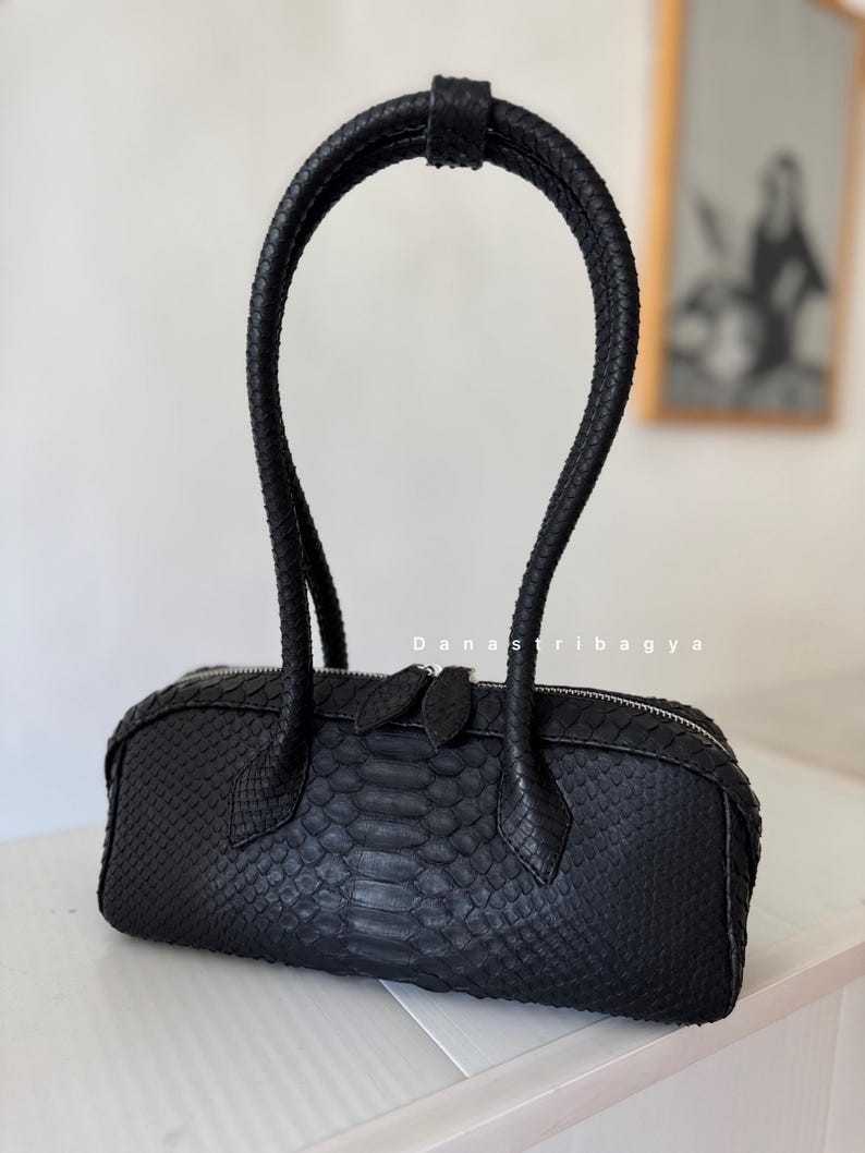 May include: A black cylindrical handbag with a textured snakeskin pattern, featuring a single tall braided handle and a silver-toned zipper closure with two pull tabs. The text Danastribagya is printed in white above the center of the bag.