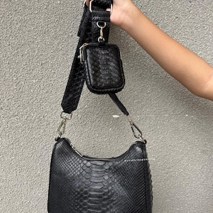 May include: Black crossbody bag with a snake-skin pattern, a small matching pouch, and a wide adjustable strap. The bag has silver-tone hardware and a zipper closure. The bag is held up by a person's hand.