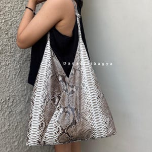 Könnte beinhalten: Eine große, beige und graue Tragetasche mit Schlangenmuster und einem einzelnen Schulterriemen. Die Tasche hat eine V-förmige Öffnung und der Text "Dana Ibagya" ist sichtbar.