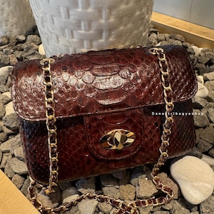 Può includere: Una borsa in pelle bordeaux con tracolla a catena dorata. La borsa ha un motivo strutturato e scaglioso e una chiusura dorata. La borsa è appoggiata su un letto di piccole pietre grigie. Il testo "DanaTribagyshop" è visibile.