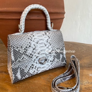May include: A small, rectangular handbag with a snakeskin pattern in shades of gray and white. It features a short, rounded handle and a detachable shoulder strap. The bag has a clasp closure and is set on a wooden surface.