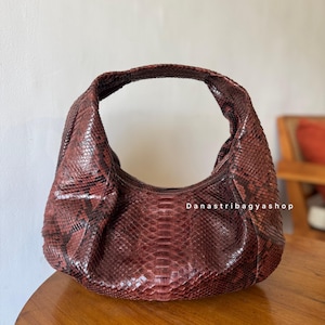 Peut inclure: Un sac à main en forme de croissant de couleur bordeaux avec un motif peau de serpent. Le sac a une seule poignée et une forme arrondie. La texture de la peau de serpent est visible, et le sac semble être en cuir. Le texte "Danastribagyashop" est visible.