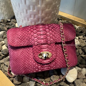 Może przedstawiać: Magenta torebka z fakturą skóry węża. Torebka posiada złoty łańcuszek i złote zapięcie. Widoczny jest napis "Danetribagyashop". Torebka jest prostokątna z klapką.