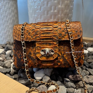 Könnte beinhalten: Kleine, rechteckige Handtasche mit Schlangenmuster in Braun- und Orangetönen. Die Tasche hat einen goldfarbenen Kettenriemen und einen Verschluss mit silbernem und goldenem Design. Die Tasche wird auf einem Bett aus grauen und weißen Steinen präsentiert.