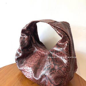 Peut inclure: Sac &agrave; bandouli&egrave;re marron et noir &agrave; motif peau de serpent, de forme incurv&eacute;e. Le sac a une seule bandouli&egrave;re et une fermeture &eacute;clair. Le texte "Danasnagyashop" est visible sur le c&ocirc;t&eacute; du sac. Le sac est pos&eacute; sur une surface en bois.
