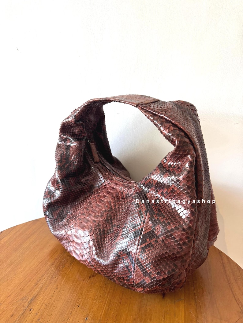 Peut inclure: Sac &agrave; bandouli&egrave;re marron et noir &agrave; motif peau de serpent, de forme incurv&eacute;e. Le sac a une seule bandouli&egrave;re et une fermeture &eacute;clair. Le texte "Danasnagyashop" est visible sur le c&ocirc;t&eacute; du sac. Le sac est pos&eacute; sur une surface en bois.