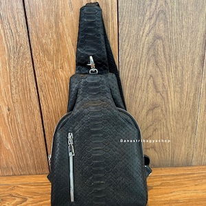 Może przedstawiać: Czarna torba na ramię z wzorem skóry węża. Posiada przednią kieszeń zapinaną na zamek błyskawiczny i srebrny zamek. Czarny pasek jest przymocowany srebrną klamrą. Widoczny jest tekst "Danastribagyashop".