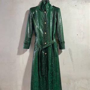 May include: A long, emerald green coat with a snakeskin pattern. The coat features a high collar, gold buttons, and a matching belt. The coat is hanging against a textured, light gray wall.
