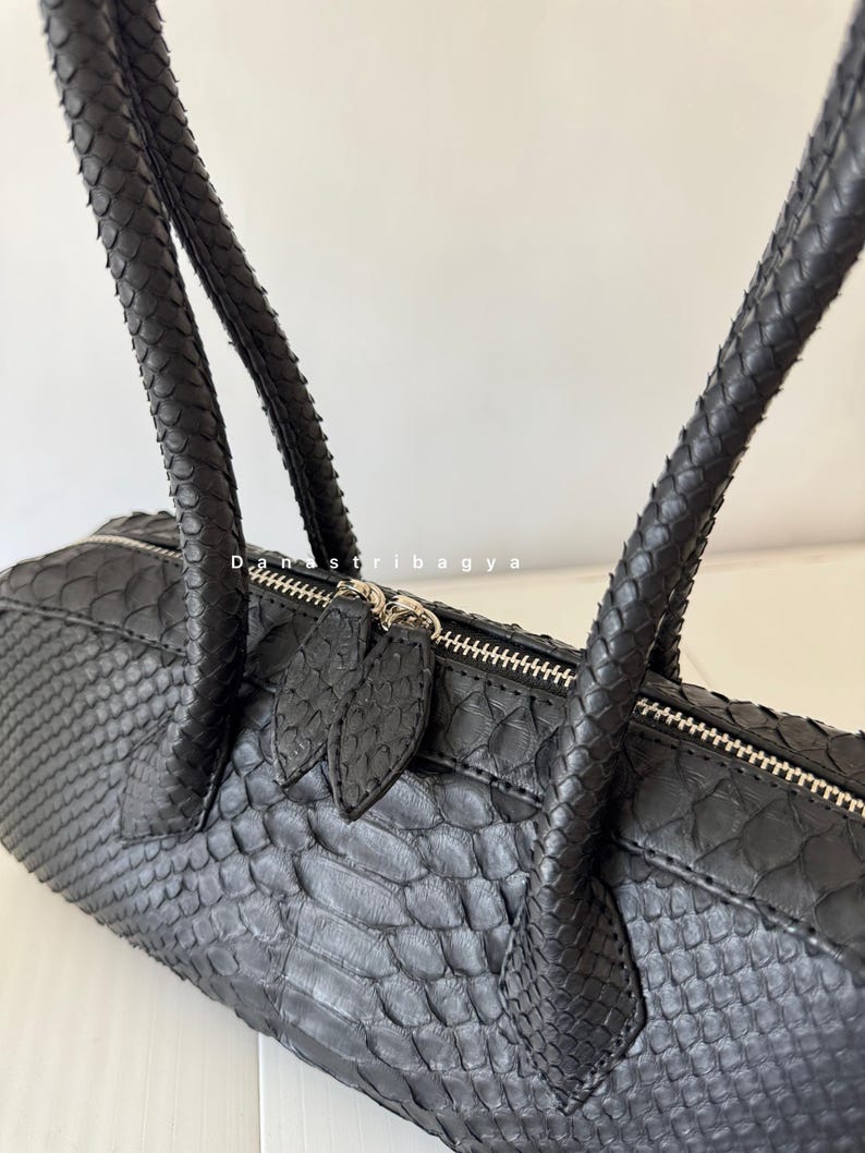 May include: A black handbag with a textured, scale-like pattern across the exterior. The bag features two matching handles and a silver-toned zipper closure with two leaf-shaped pulls. The text "Danastribagya" is printed in white near the zipper.