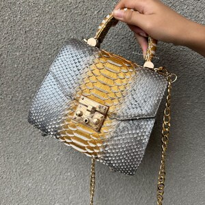 Puede incluir: Un bolso de mano plateado y dorado con un patrón de piel de serpiente texturizado. El bolso tiene una correa de cadena dorada y un cierre de broche dorado. El asa también es dorada y tiene un patrón de piel de serpiente.