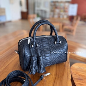 Può includere: Borsa a mano in pelle nera con motivo a pelle di serpente, con due manici intrecciati e una tracolla staccabile. La borsa ha una chiusura a zip e nappe decorative. Il testo "DanaStribagyshop" è visibile.