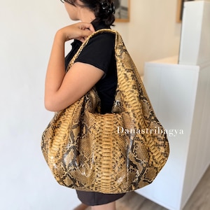 May include: A large hobo-style shoulder bag featuring a textured snakeskin pattern in shades of golden brown, tan, and dark brown. The bag has a slouchy, unstructured silhouette with a wide shoulder strap and the text Danastribagya printed on it.