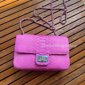 以下が含まれることがあります： 明るいピンク色のハンドバッグ。蛇革のような質感で、シルバーのチェーンストラップとシルバーとブルーの留め金が付いています。「Danastribagyashop」の文字が見えます。