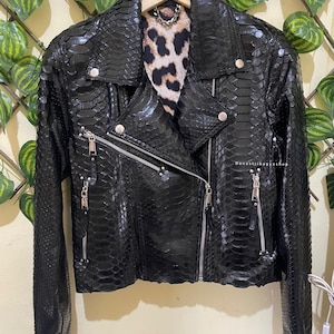 Może przedstawiać: Czarna skórzana kurtka motocyklowa z efektem skóry węża. Posiada asymetryczny zamek błyskawiczny, srebrne okucia i podszewkę w panterkę. Kurtka wisi na drewnianym wieszaku. Klasyczny kołnierz z klapami.