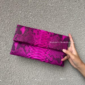 Puede incluir: Un bolso de mano magenta vibrante con un diseño de piel de serpiente. El bolso rectangular tiene una solapa plegable. El texto "Danastribagyashop" es visible en el lado derecho.