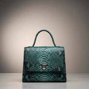 Op de afbeelding: Een teal groene handtas met een slangenleerpatroon. De tas heeft een gestructureerde vorm met een handvat aan de bovenkant en een zilverkleurige sluiting. De tas staat op een lichtgekleurd oppervlak.