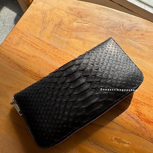 May include: A black, rectangular wallet with a textured, scaly pattern, likely made of leather. The wallet has a zipper closure and is resting on a wooden surface. The text "Danastribagyashop" is visible on the wallet.