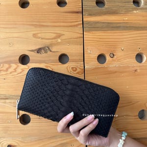 May include: A black, textured wallet with a zipper closure. The wallet is held against a wooden background. The texture of the wallet appears to be snake-skin like. The text "DanaTribagyshop" is visible on the wallet.