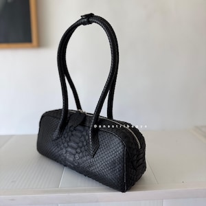 May include: A black cylindrical handbag with a textured snakeskin pattern, featuring two tall, rounded handles and a silver-toned zipper closure along the top. The text "danastribagya" is printed in small white letters on the side of the bag.